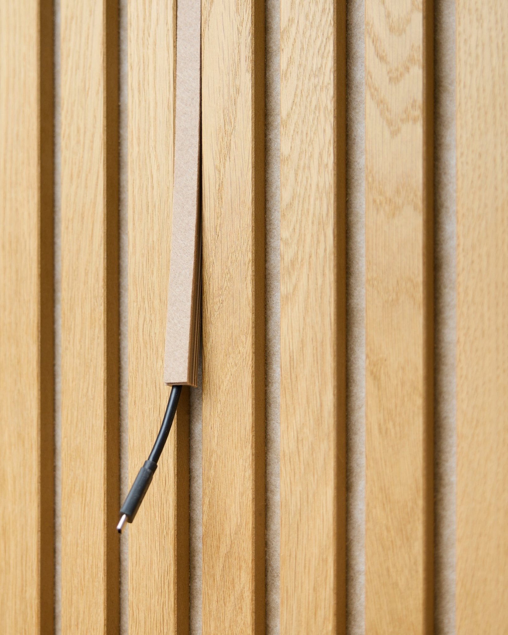 Un cable black con gestión de cables queda parcialmente oculto en un Dupps Beige Pop-on Cable Cover de WoodUpp, montado sobre lamas acústicas de madera vertical Akupanel.